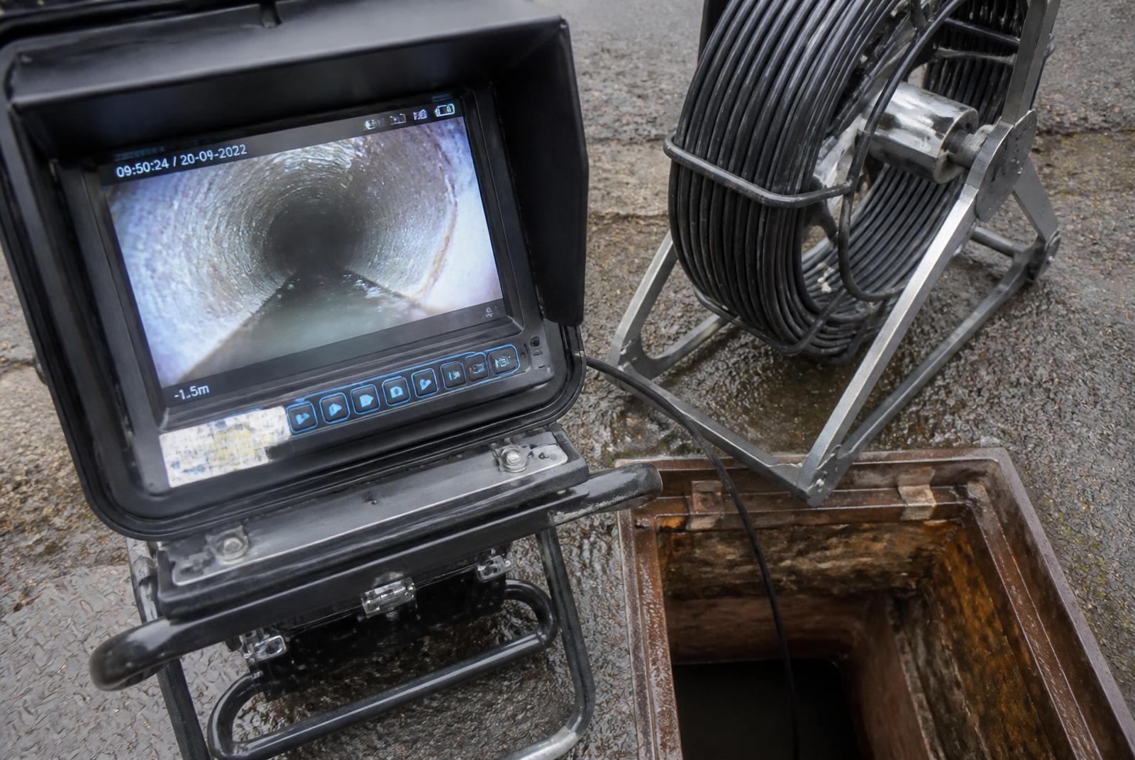 Drain survey monitor displaying live camera footage from inside a drainage pipe, next to a camera reel and open chamber